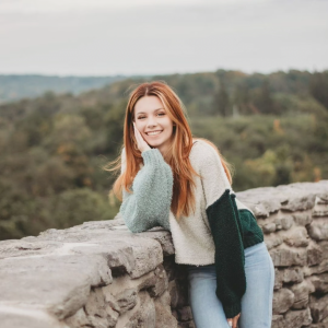 A young woman with long, red-brown hair leans against a stone wall