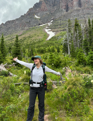  A smiling person with outstretched arms standing in a forest with a mountain in the background. 