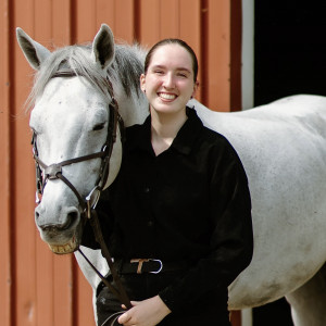 Pictured is a figure in all black standing next to a white horse. 